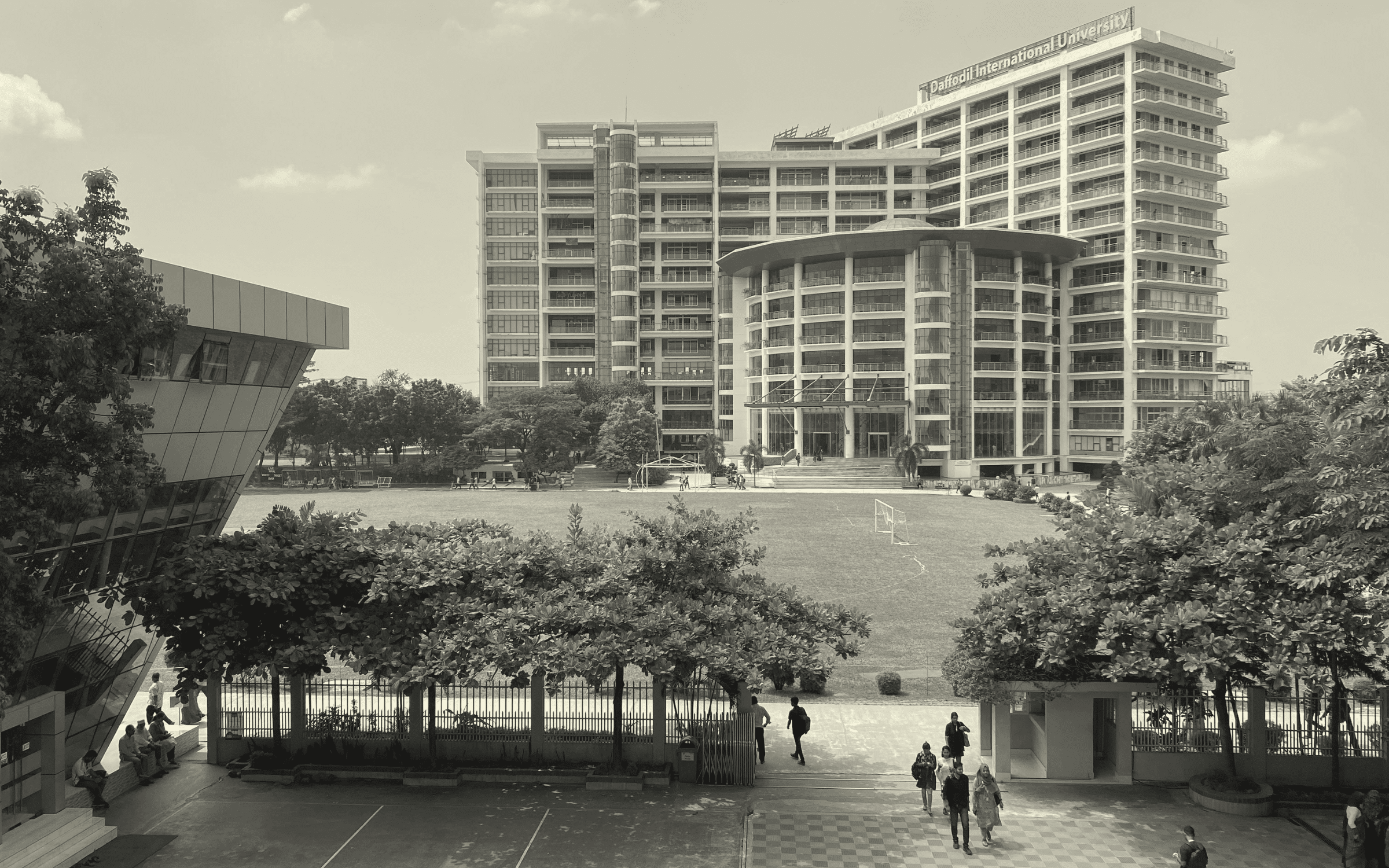 Wide view of a university campus with a large modern building, trees, and a sports court in the foreground.